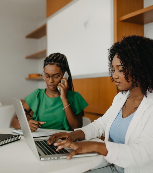 Two women are sitting at a table. The woman in the front is typing on a laptop, while the woman in the back is on the phone and taking notes.