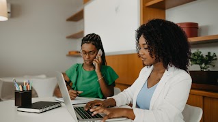Two women are sitting at a table. The woman in the front is typing on a laptop, while the woman in the back is on the phone and taking notes.