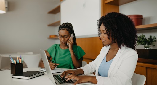 Two women are sitting at a table. The woman in the front is typing on a laptop, while the woman in the back is on the phone and taking notes.