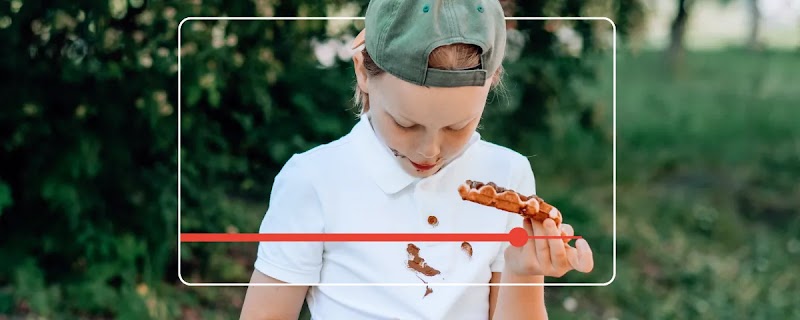 Inside a browser screen icon, a child wearing a baseball cap backwards is eating a chocolate waffle. His white polo shirt is stained with chocolate. The child is standing on a patch of grass, in front of a bush.