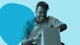 A bearded Black man wearing glasses sits cross-legged while enthusiastically opening a package he’s received.