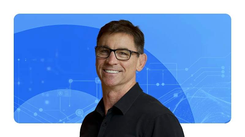 Paul Limbrey, Google’s VP of global 3P ecosystem, appears in front of a background of blue arcs. Limbrey has light skin, dark hair, and wears glasses and a black collared shirt.