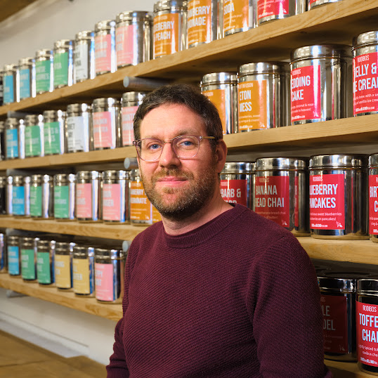 صورة لرجل بشعر بني قصير ولحية ونظارات يرتدي سترة كنزة بلون ماروني، يقف أمام عدة أرفف خشبية مليئة بعبوات شاي فضية كبيرة. تتميز كل عبوة بملصق ملون بنكهات شاي متنوعة، مما يخلق خلفية حيوية ومنظمة في متجر شاي أو مستودع.