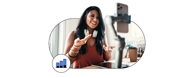 Una mujer sonríe a una videollamada en su teléfono móvil, sentada a una mesa. Pelo oscuro, gafas, auriculares y blusa rosa.