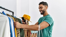 A happy arabian man with a beard and a green t-shirt is holding a phone and a white shopping bag while browsing a rack of clothes in a store.