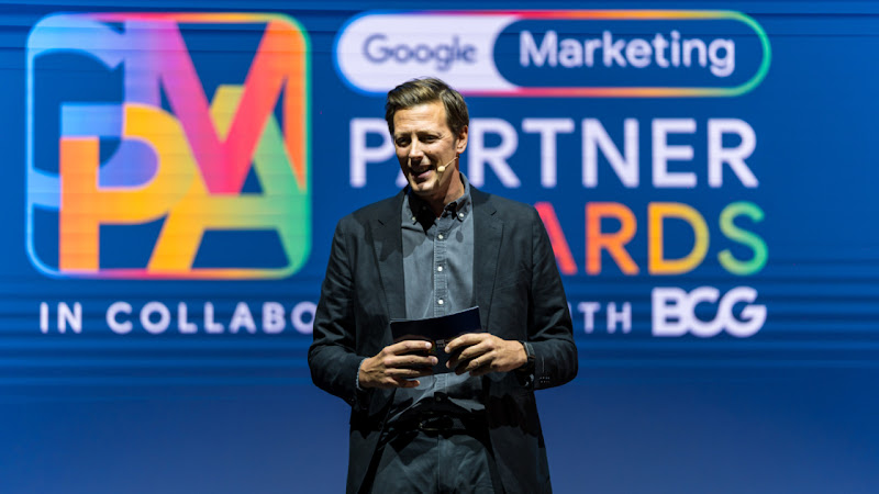 Harry Walker de medio cuerpo con traje de chaqueta oscuro y camisa gris sujeta unas tarjetas mientras habla sobre el escenario de los GMPA, el fondo es azul y se ve el logo y las letras de colores de los Google Marketing Partners Awards.