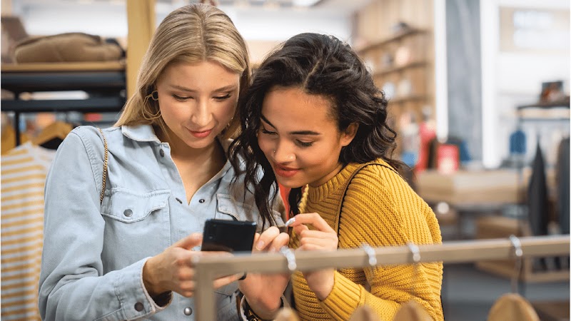 Inside a shop, two women are looking intently at a mobile phone screen. One has long blonde hair, and one has long dark hair. The woman with blonde hair is holding the phone. Visible in the background are clothing items on hangers.