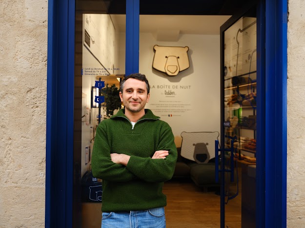 Un homme souriant aux cheveux bruns et courts et à la barbe naissante pose les bras croisés à l'entrée bleue et ouverte d'une boutique. Il porte un pull en maille côtelée vert foncé à col zippé et un jean bleu. En arrière-plan, à l'intérieur du magasin, un logo en bois en forme de tête d'ours est accroché au mur au-dessus de l'inscription « LA BOÎTE DE NUIT by Tediber », accompagné d'articles de literie et de coussins à motif d'ours de la marque.