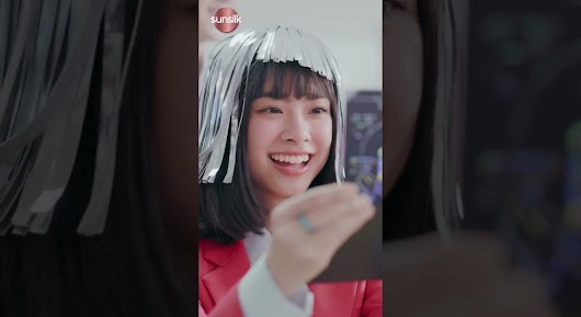 A vertical video thumbnail of a smiling young woman in a red blazer, with strange silver strips stuck to her hair, holding out a small blue card.