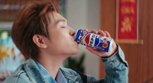 A close-up profile shot of a young man with light brown hair wearing a light blue denim jacket. He has his eyes closed as he takes a drink from a blue Pepsi can featuring a special festive design. In the blurred background, there is a traditional red and gold framed decoration on a wooden wall, along with some indistinct plants.