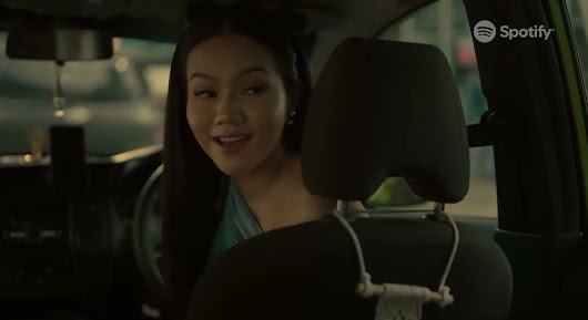 A video screenshot of a smiling woman looking back from the driver's seat of a car, with the Spotify logo in the top right corner.