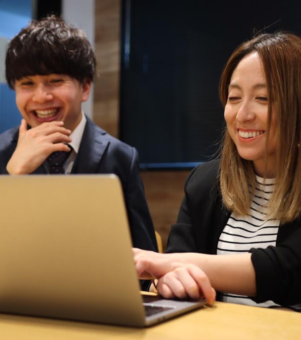 A man and a woman wearing office attire are sitting at a table and looking at a laptop, smiling. There is a black screen behind them.