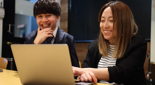 A man and a woman wearing office attire are sitting at a table and looking at a laptop, smiling. There is a black screen behind them.