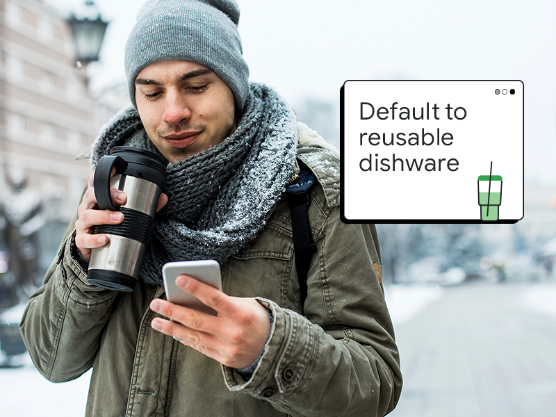 A man with medium skin wearing winter outerwear and accessories carries an insulated coffee mug while using a mobile device as he walks down a snowy street. A pop-up window reads, “Default to resuable dishware.”