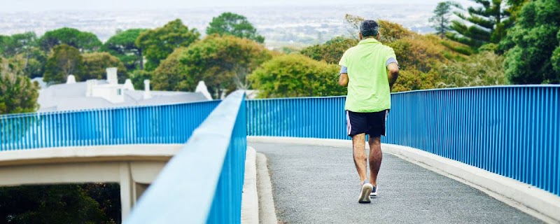 Un uomo in abbigliamento sportivo fa jogging su un ponte con ringhiere blu, circondato da alberi.