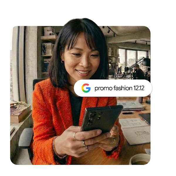 Seorang wanita berjaket oranye tersenyum sambil melihat ponsel pintar di meja kerjanya, dengan kolom pencarian Google bertuliskan "promo fashion 12.12" di depannya.