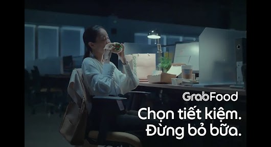 The video thumbnail shows a group of co-workers looking stressed or busy in an office environment, contrasting with food, to promote saving money with GrabFood.