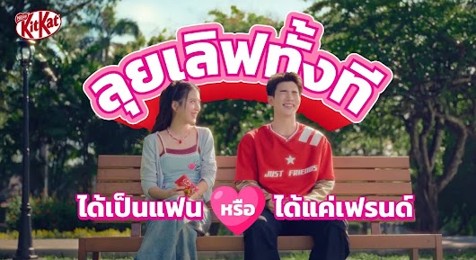 A KitKat video thumbnail showing a smiling young man and woman sitting on a park bench, accompanied by pink Thai text about romance.