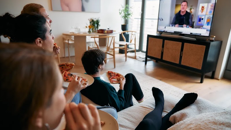 Family sitting together on a living room couch, eating slices of pizza while watching a man present on a large television screen. 