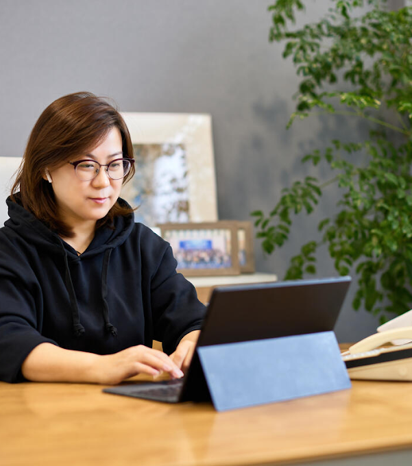 A woman wearing glasses and sitting at a desk is typing on a laptop. There is a mobile phone as well as a desktop phone on the table.