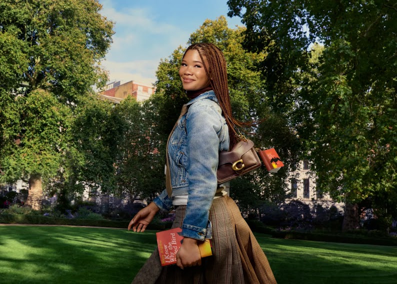 A young woman with brown skin and long braided hair carries a book across a leafy campus. She wears a blue denim jacket with a plaid pleated skirt, and carries a Coach Tabby bag with a book charm over her shoulder.