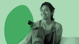 Uma mulher usando um macacão e uma camiseta está sentada em um sofá, sorrindo e apontando um controle remoto de TV.