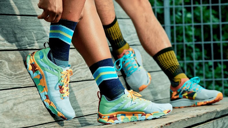 Two people, wearing Smartwool socks and athletic shoes, stand on a wooden bench. Only their legs, from the knee down, are in frame. The hands of the foreground person reaches down to adjust their black-and-aqua striped Smartwool socks.