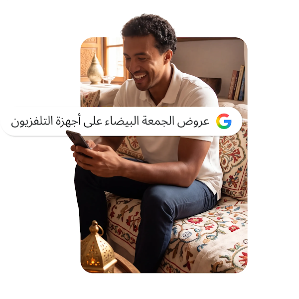 صورة لرجل مبتسم يجلس على أريكة، يستخدم هاتفًا ذكيًا مع تراكب شريط بحث Google مكتوب عليه "عروض الجمعة البيضاء على التلفزيون".