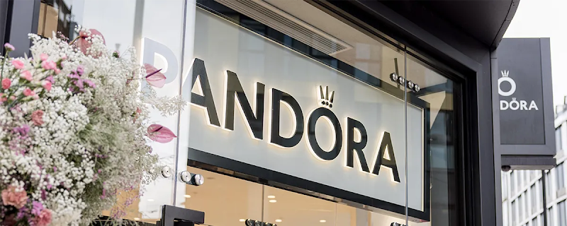 A Pandora store entrance featuring an illuminated storefront sign reading "PANDORA" with a crown logo over the letter O, framed by soft pink and white flowers on the left and a black exterior blade sign on the right.