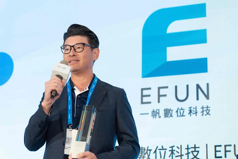一帆數位科技人員於Google頒獎典禮上登台演講，手持麥克風。講者著深藍色西裝、白色襯衫，佩戴藍色證件帶與眼鏡。背景展示一帆數位科技(EFUN)品牌大藍色標誌，舞台燈光呈冷調藍白色。此演講呈現該公司導入Google AI驅動的金融市場策略成果。