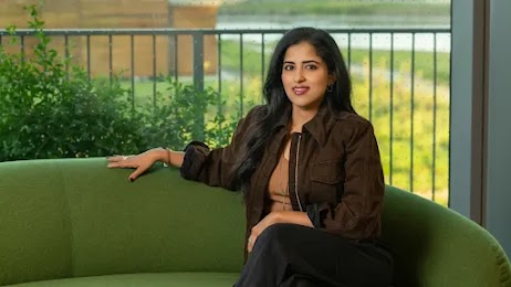 Vidhya Srinivasan, vice-présidente et directrice générale Ads and Commerce chez Google, est assise sur un canapé vert devant une fenêtre donnant sur un arbuste et une rambarde de terrasse. Elle a la peau mate, de longs cheveux foncés et porte une veste marron décontractée sur un haut beige et un pantalon noir.