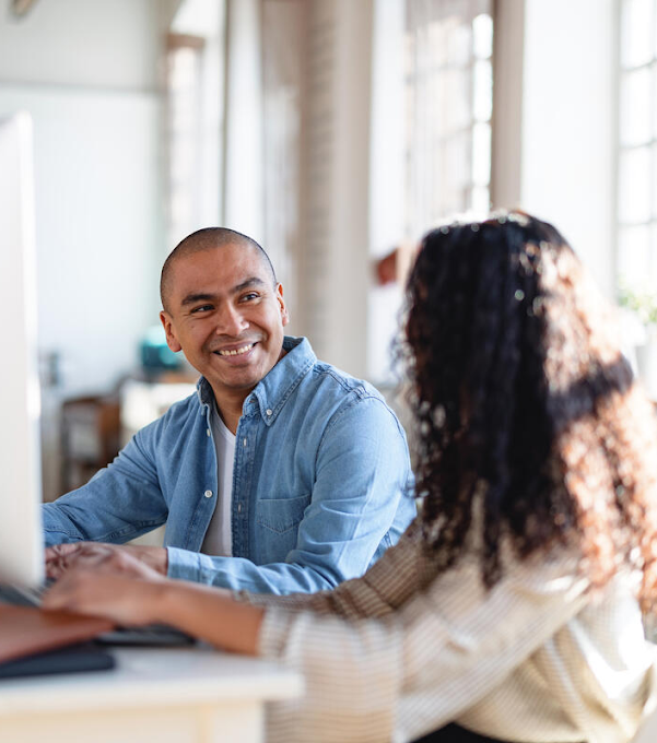 A man and a woman are sitting at a desk in front of a desktop computer. The man is smiling and the woman is turned away, her face hidden.