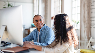 A man and a woman are sitting at a desk in front of a desktop computer. The man is smiling and the woman is turned away, her face hidden.
