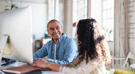 A man and a woman are sitting at a desk in front of a desktop computer. The man is smiling and the woman is turned away, her face hidden.