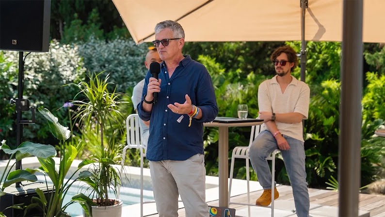 Google DeepMind’s Doug Eck holds a microphone and speaks from a patio under large shading umbrellas. Eck has light skin and short silver hair, and wears sunglasses and a dark blue button-front shirt. Two men sit behind him at high-top tables.