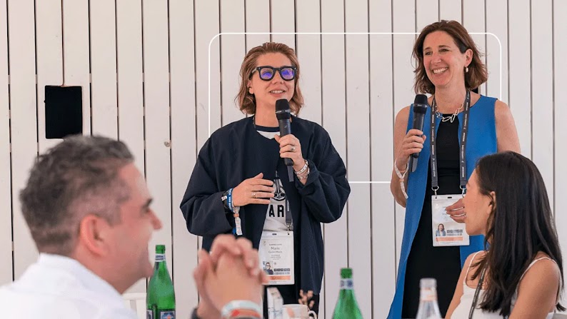 Marie Gulin-Merle y Alison Wagonfeld, VPs de Google, hablan ante un grupo de invitados del Cannes Lions que se encuentran sentados alrededor de las mesas. Las mesas están llenas de botellas de agua con gas y flores secas. Todos ríen y sonríen.