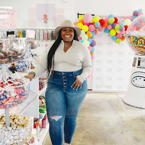 The business owner of Cypress Sweets standing and posing for a photo in her sweet shop, wearing casual clothes and a hat.