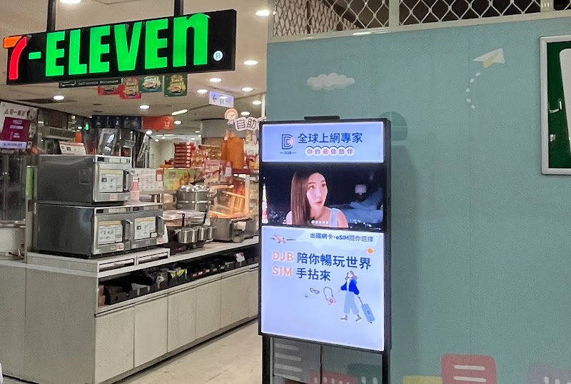 7-ELEVEN 綠紅橘招牌門市入口處，兩位行人經過櫥窗旁的直立式數位看板，畫面播放品牌人物廣告，由 Google Ads 與 Google Marketing Platform 透過 DV360 程序化廣告同步投放，串連 DOOH 與線上轉換路徑。