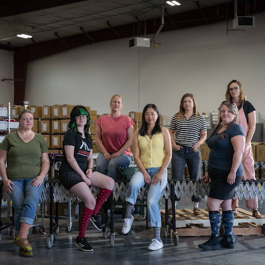 The Vim & Vigr team stand together in their warehouse with a conveyor belt behind them. 