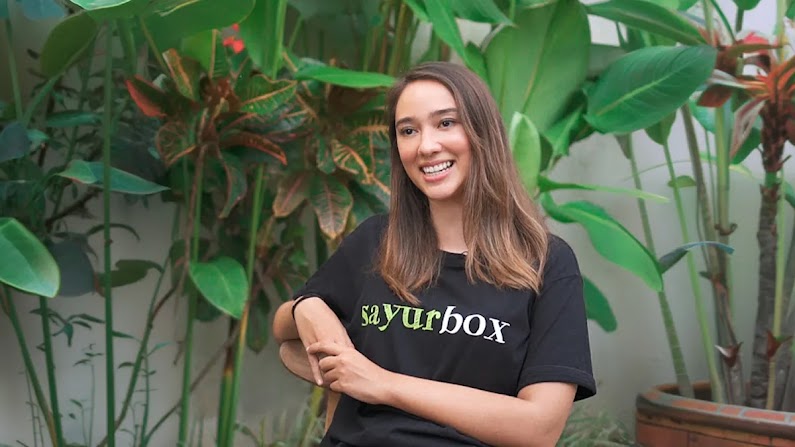 Seorang wanita mengenakan kaos berlogo Sayurbox, tersenyum ke arah kamera