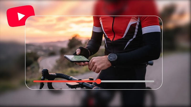 A male cyclist in an orange and black top and wearing a watch sits on his bike, holding a phone. The outer edges are blurred, and a YouTube play icon and progress bar is overlaid in the centre.