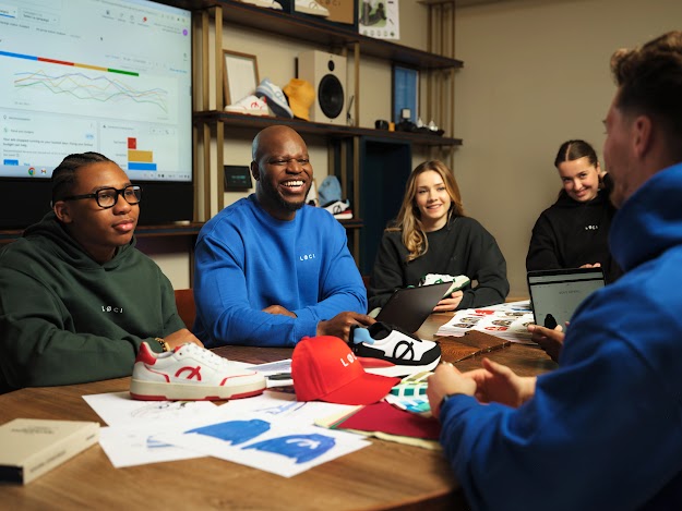  A team of people collaborate around a wooden table filled with sneakers, design sketches, and a red cap. In the background, a large digital screen displays a Google Ads dashboard showing performance graphs, while shelves behind them showcase various footwear and apparel samples.