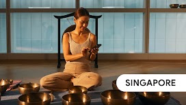 Singapore. A woman sitting cross-legged on the studio floor using her mobile phone, with many Tibetan singing bowls before her and a gong behind her.
