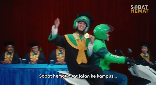 The video thumbnail shows a student in a graduation cap and gown, wearing a helmet and green headphones, riding on the back of a Grab driver's motorbike. The student is holding up a gold medal or disc while smiling excitedly. The scene takes place indoors with a red curtain backdrop and several people dressed in graduation robes seated at a table in the background. The text overlay reads: "Sobat hemat buat jalan ke kampus..." (A budget friend for the trip to campus...).