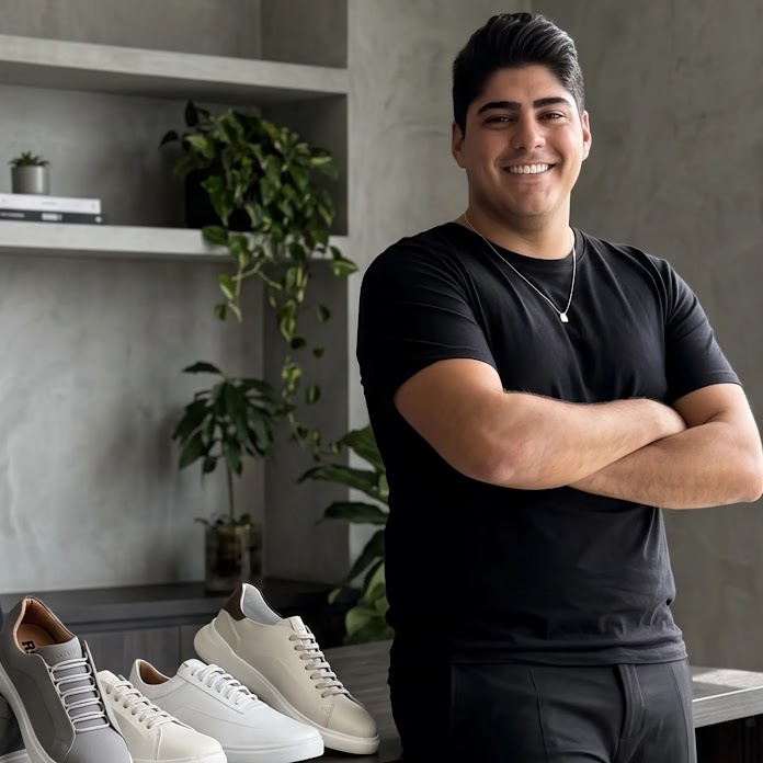 Profissional sorrindo, Guilherme Cintra, CEO da Reise, apoiado em uma mesa de escritório moderna com tênis esportivos expostos.