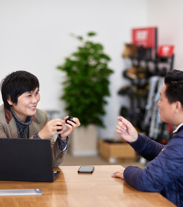 A woman and a man are sitting at a table, smiling and talking. There is an open laptop on the table, as well as a phone.