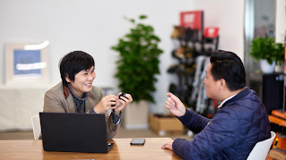 A woman and a man are sitting at a table, smiling and talking. There is an open laptop on the table, as well as a phone.
