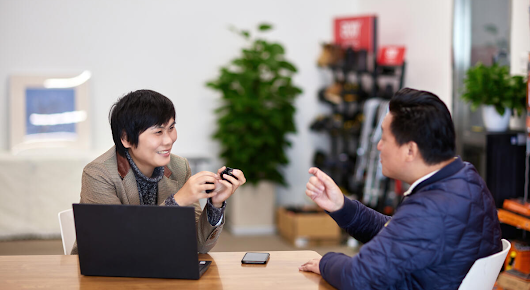 A woman and a man are sitting at a table, smiling and talking. There is an open laptop on the table, as well as a phone.