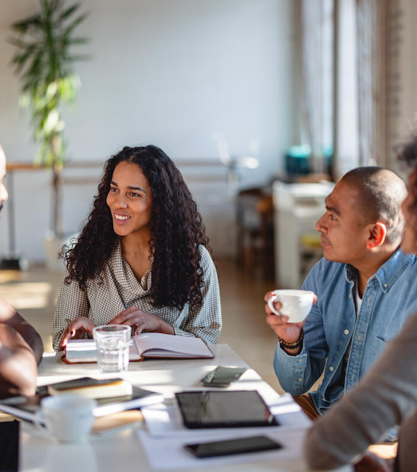 Two women and two men sitting around a table, visibly having a meeting. There are notebooks, tablets and coffee cups on the table.
