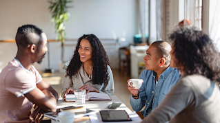 Two women and two men sitting around a table, visibly having a meeting. There are notebooks, tablets and coffee cups on the table.